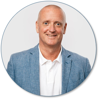 Portrait of a middle-aged man with a bald head, smiling, wearing a light blue blazer and a white shirt against a plain background.