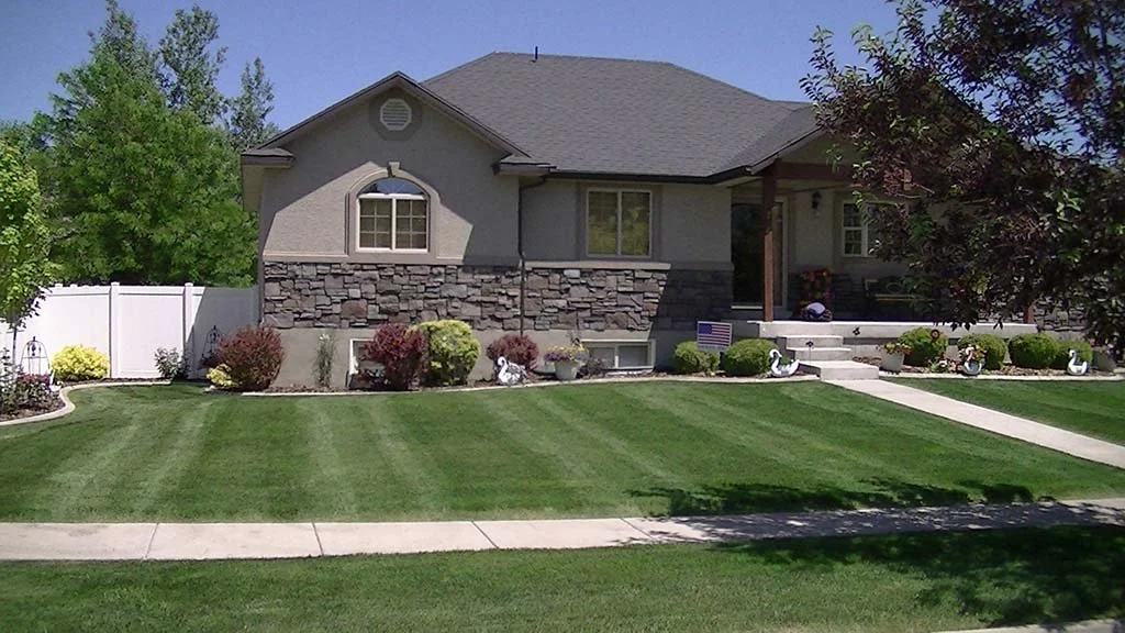 Front view of a suburban house with a well-maintained lawn, bushes, small trees, decorative swan statues, and a porch with stairs, American flag, and garden decorations.
