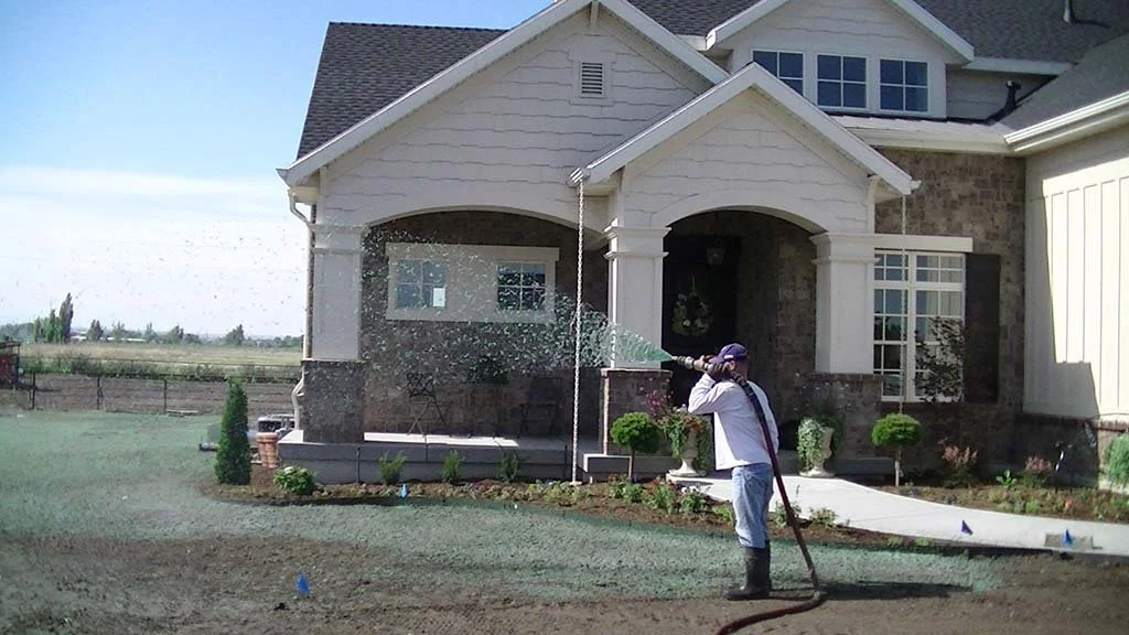 A person watering or spraying water on the front lawn of a large house using a hose.