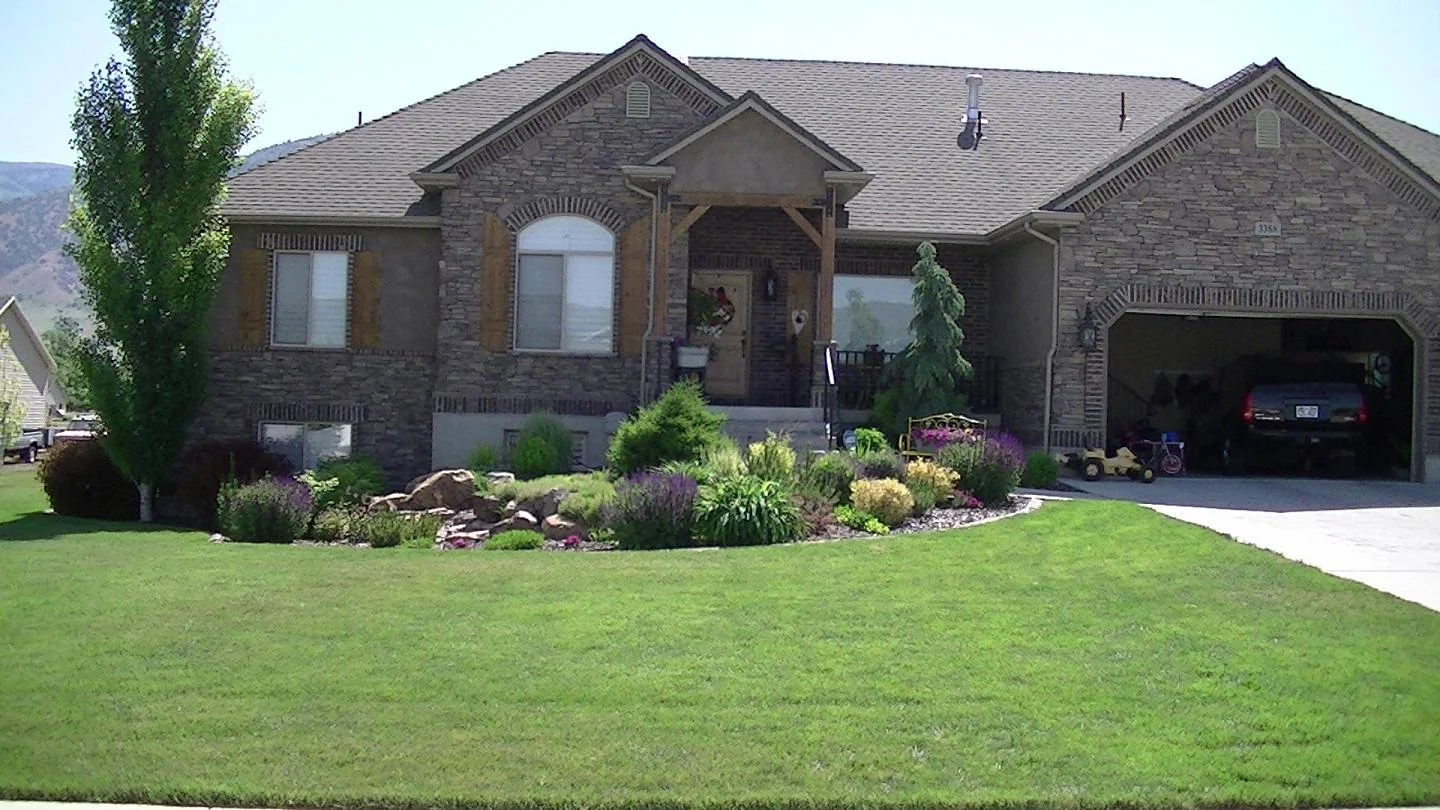 A suburban house with brick and stone exterior, a well-maintained lawn, landscaped garden, and a driveway leading to a garage.