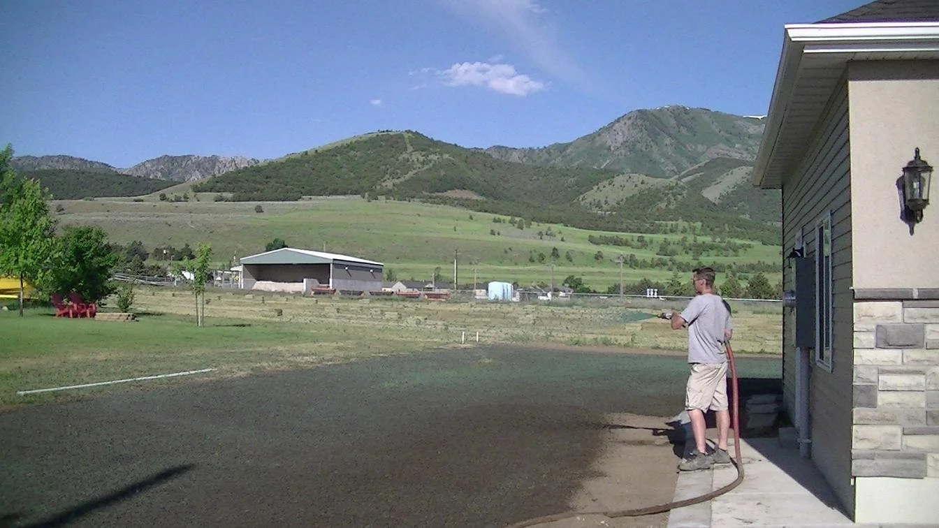 A man watering a new lawn in front of a house with mountains in the background.