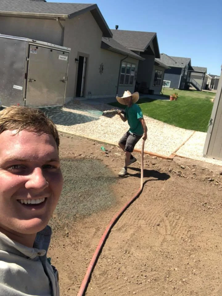 Two men are working on a backyard project. One man is taking a selfie and smiling, while the other is watering freshly poured concrete with a hose. The background shows suburban houses with green lawns and a clear blue sky.