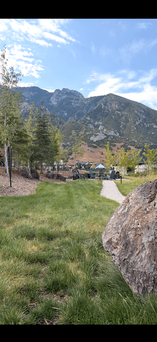 Scenic outdoor view of a mountain landscape with green trees, a pathway, patio furniture, and a clear blue sky.