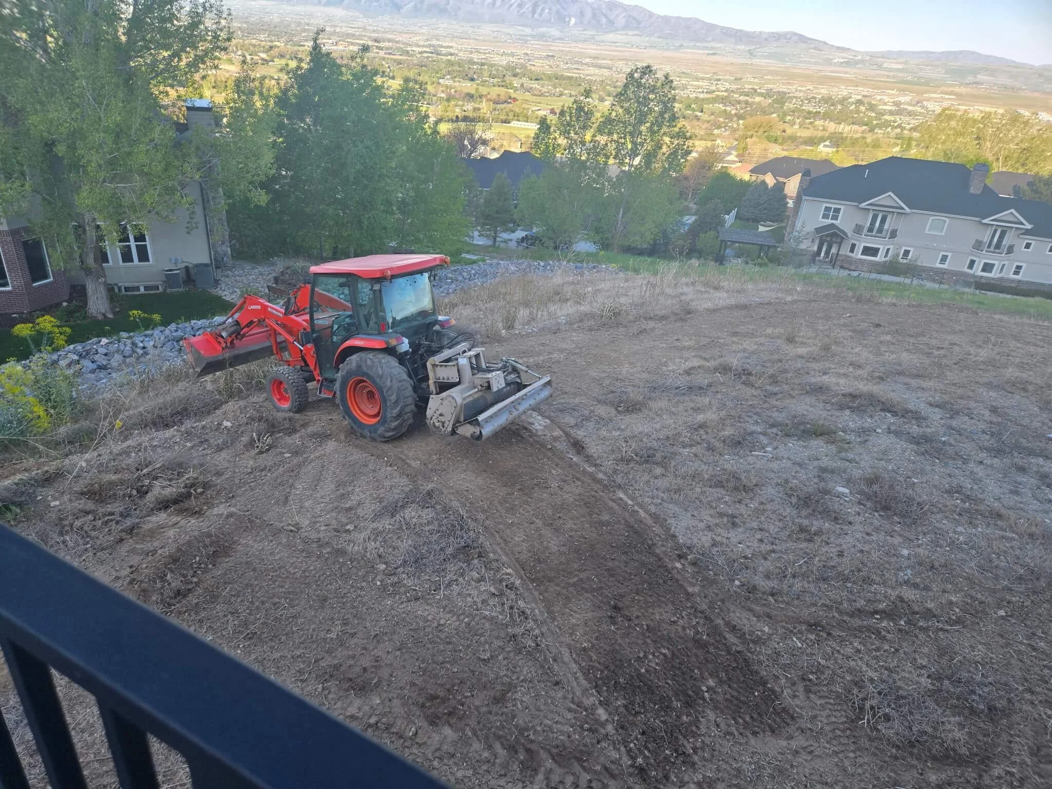 A construction site on a hillside with a red compact tractor, dirt, and rocks. In the background, there are trees, residential houses, and a distant cityscape with mountains on the horizon.
