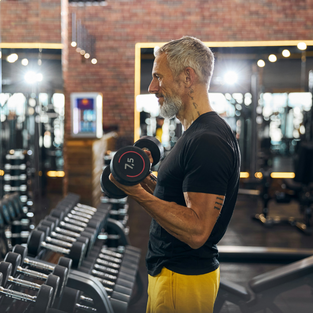 Middle-aged man with gray hair and beard lifting a dumbbell in a gym, surrounded by rows of weight racks and workout equipment.