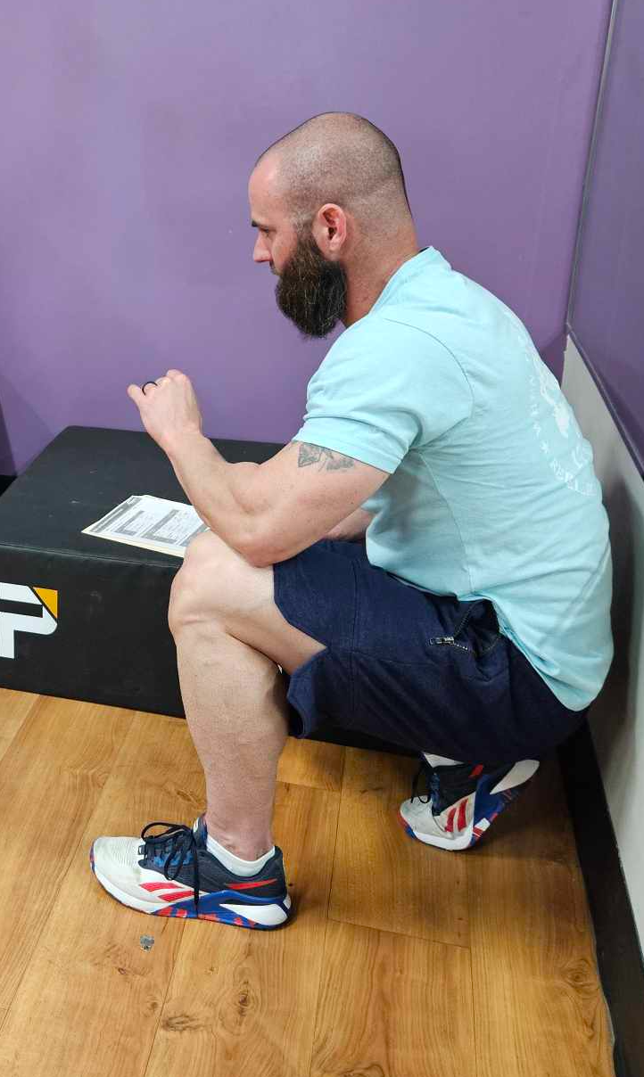 Man with bald head and full beard sitting on a bench, flexing his right arm, wearing a light blue T-shirt, dark shorts, and running shoes, with a purple wall behind him.