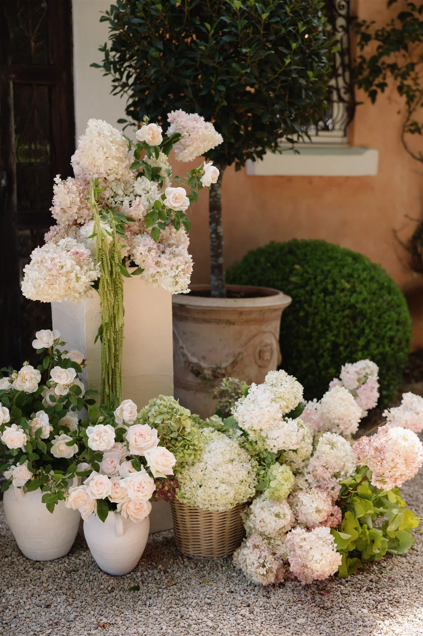 Collection of white and light pink hydrangeas and roses in baskets and vases outdoors, with potted trees and a pink wall in the background at wedding venue redleaf wollombi
