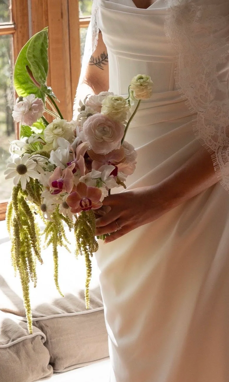 A bride holding a bouquet of white and pink flowers, standing indoors near a window with wooden framing.