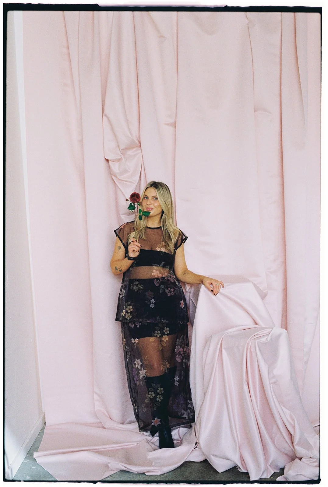 A wedding and events florist, planner and stylist standing in front of a pink fabric-draped wall, holding a rose advertising her flowers and styling business for weddings, baby showers, engagement parties, hospitality events and corporate events