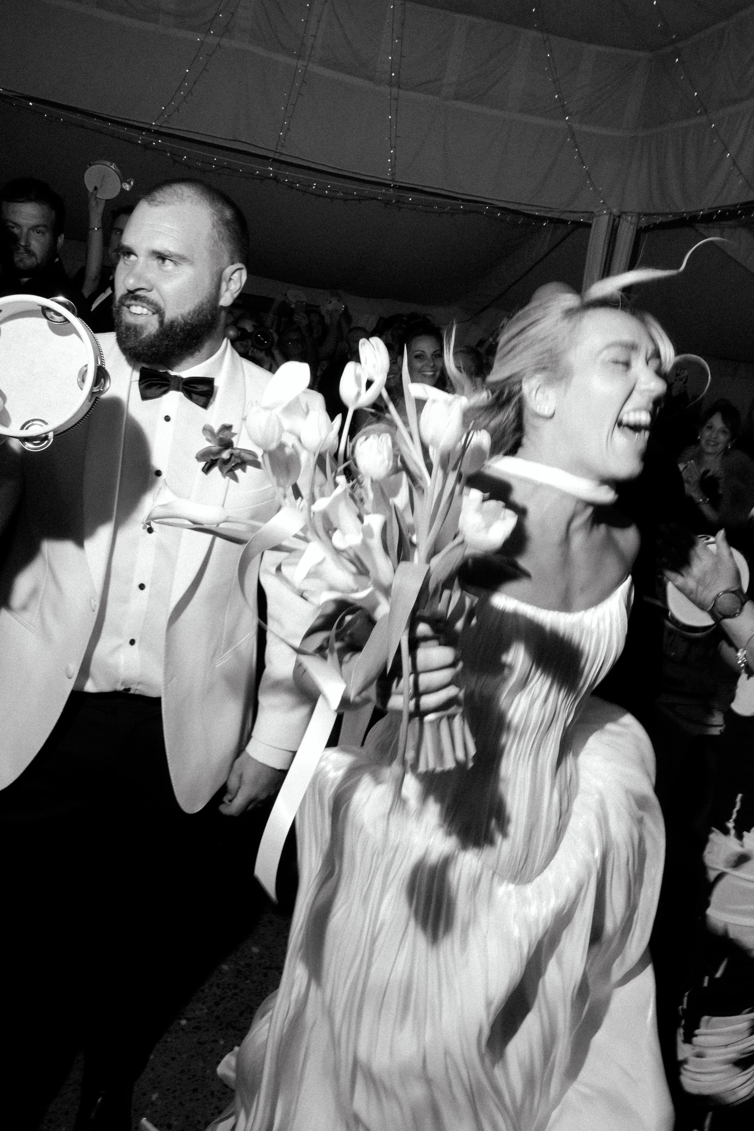 Bride and groom celebrating at a WEDDING, with a woman holding a bouquet of flowers and laughing, and a man in a tuxedo holding a tambourine, surrounded by smiling guests under string lights at Hunter Valley wedding venue Wallalong house