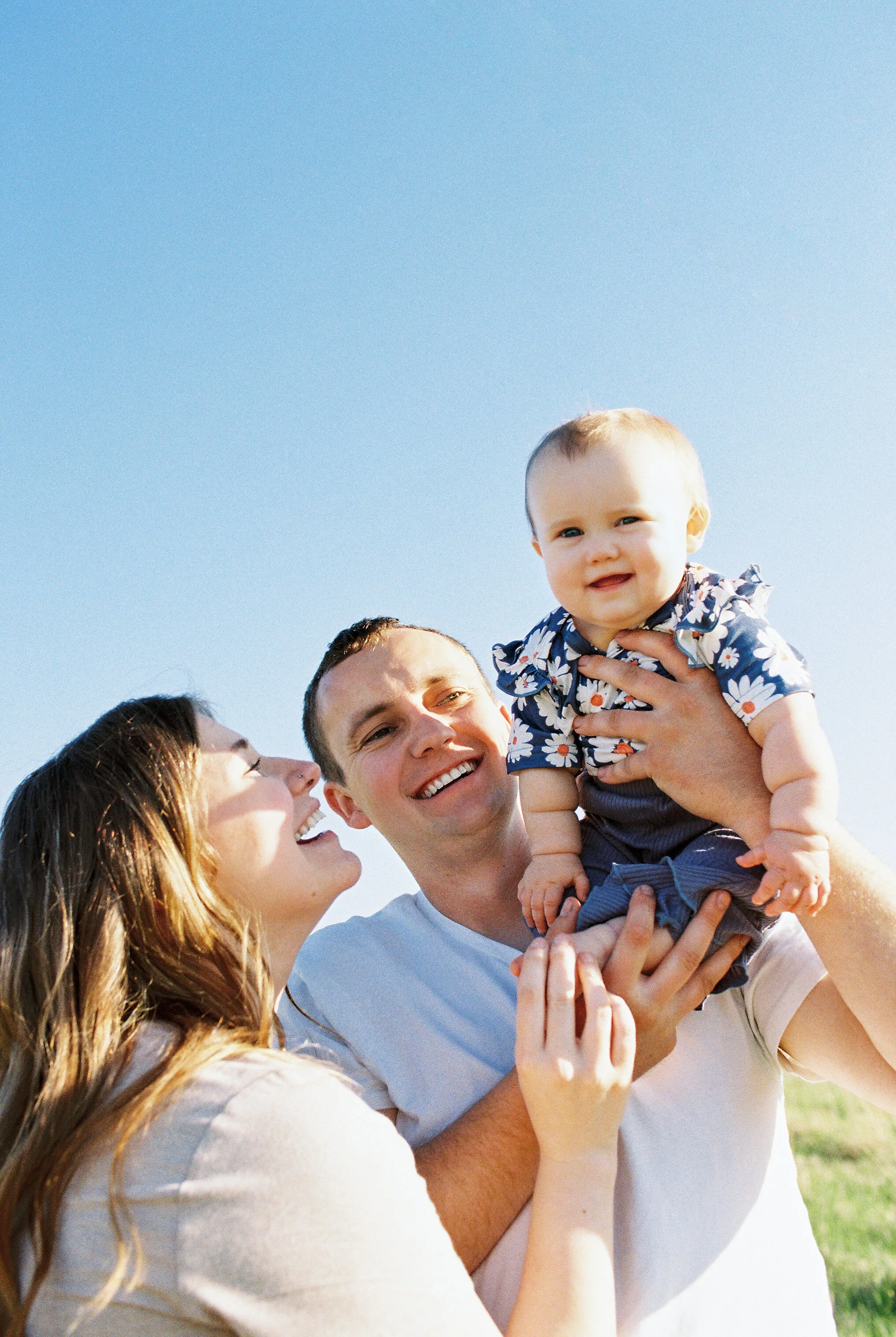 A family in a sunny field laughing together, a mother and dad holding their baby.