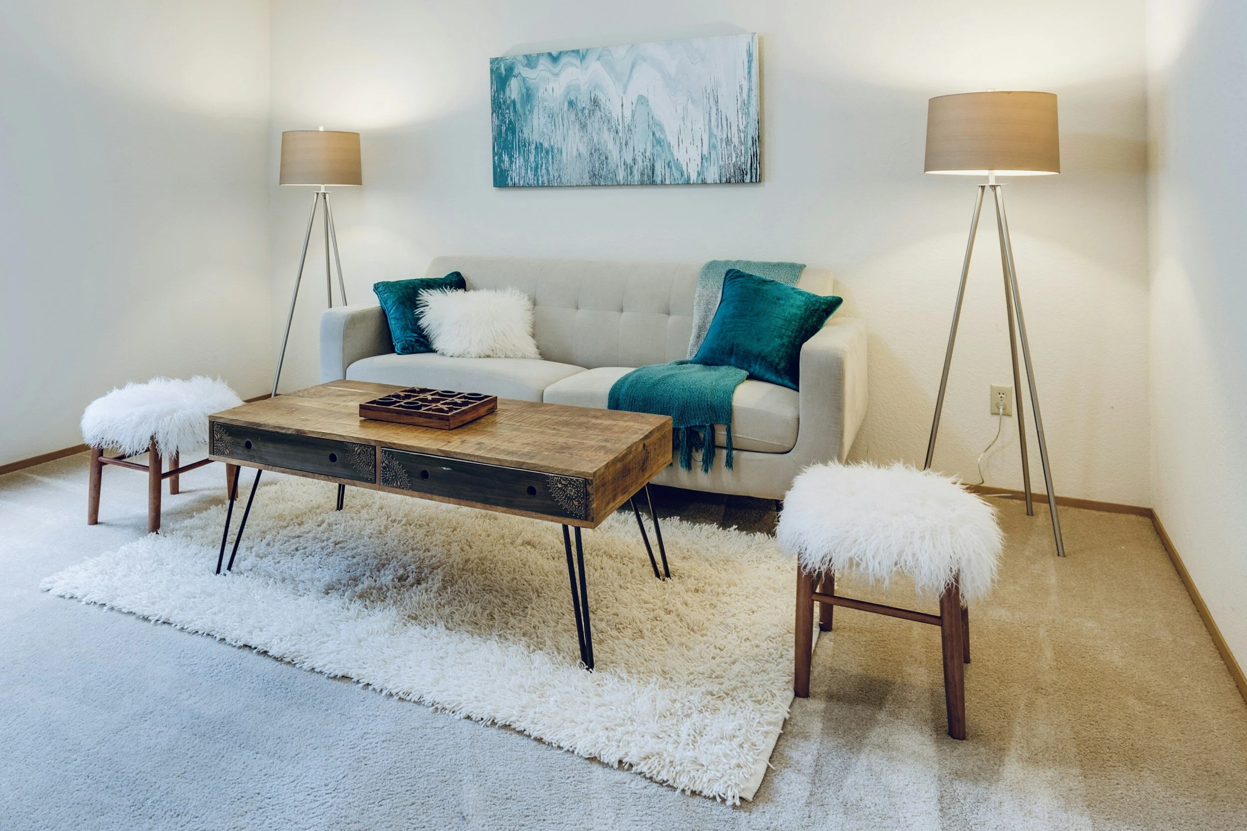 Living room with a beige sofa, blue pillows, and a throw blanket, wooden coffee table, two white furry stools, and two tall floor lamps, with a blue abstract painting on the wall.