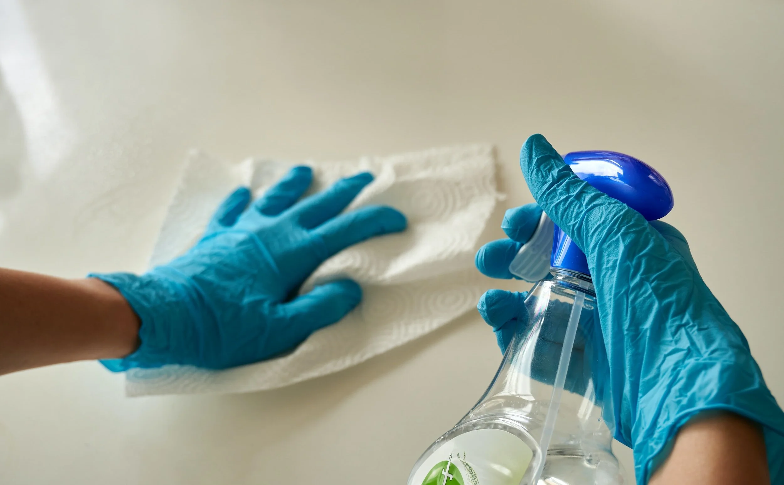 Person wearing blue gloves cleaning a surface with a paper towel while holding a spray bottle of cleaning solution.