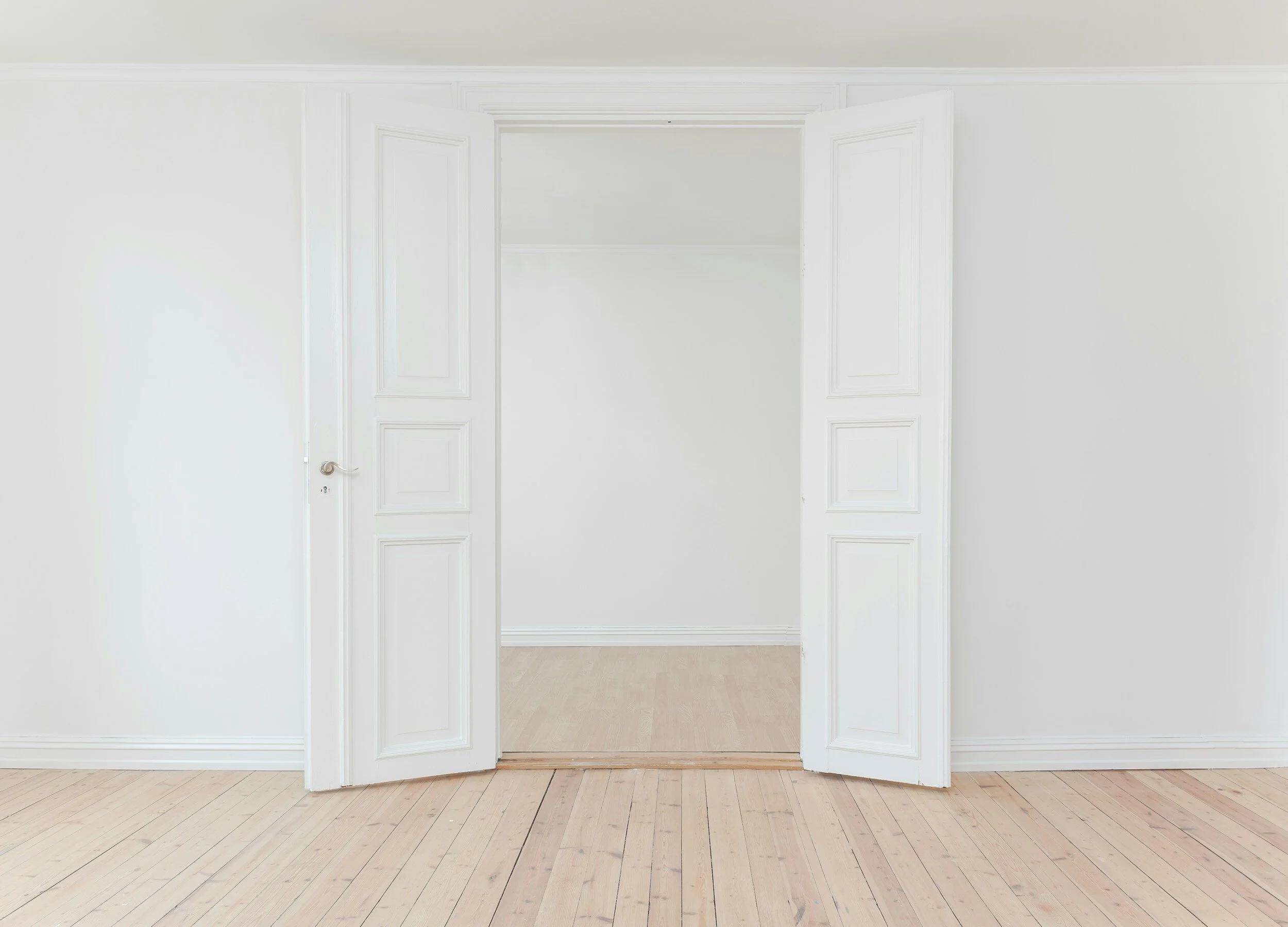 Picture of an empty room with clean white walls and tall white doors and wooden floors.