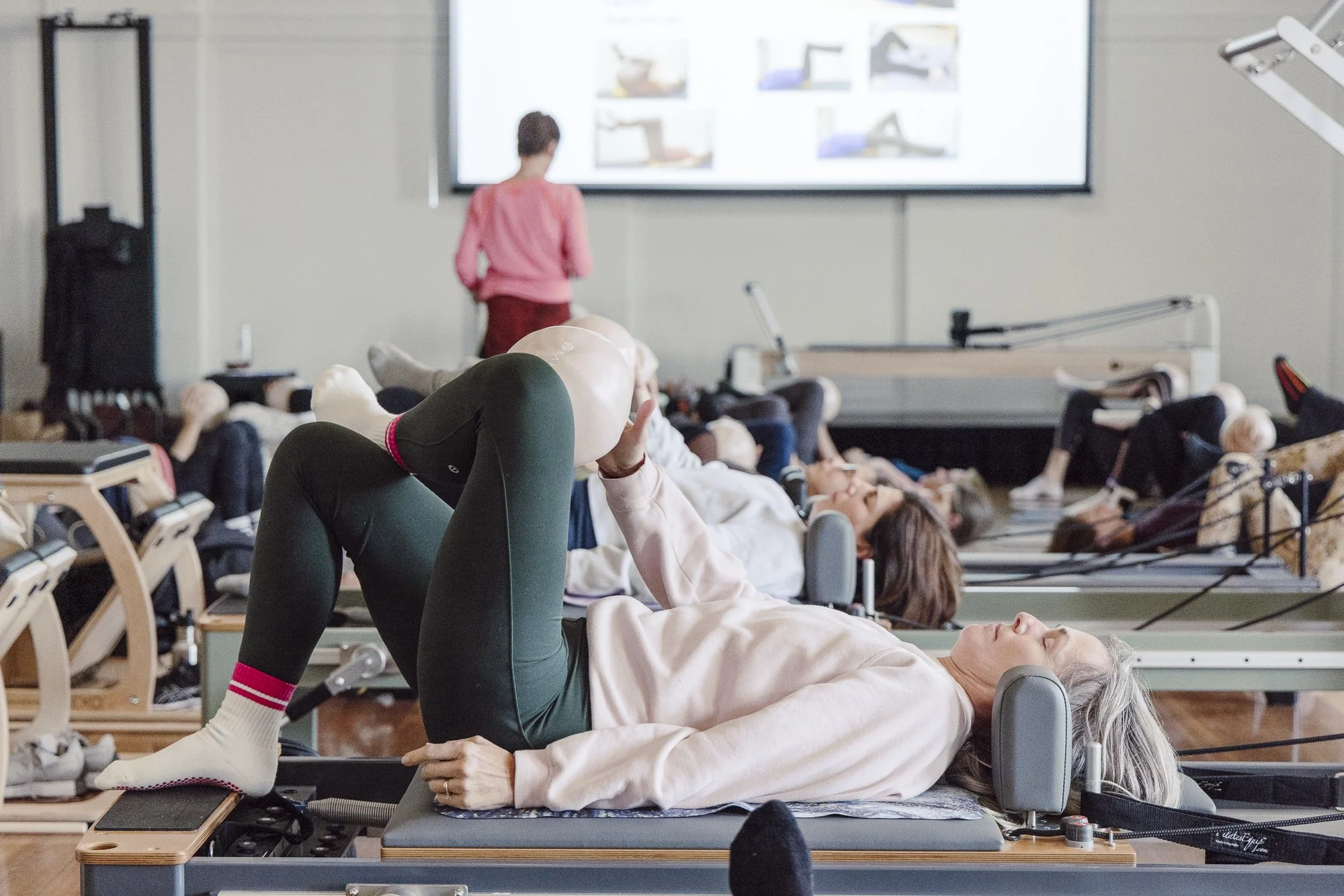 Senior woman practicing Pilates on reformer machine at The Pilates Journal Expo, with a teacher at the front and other participants on reformer machines in the background.