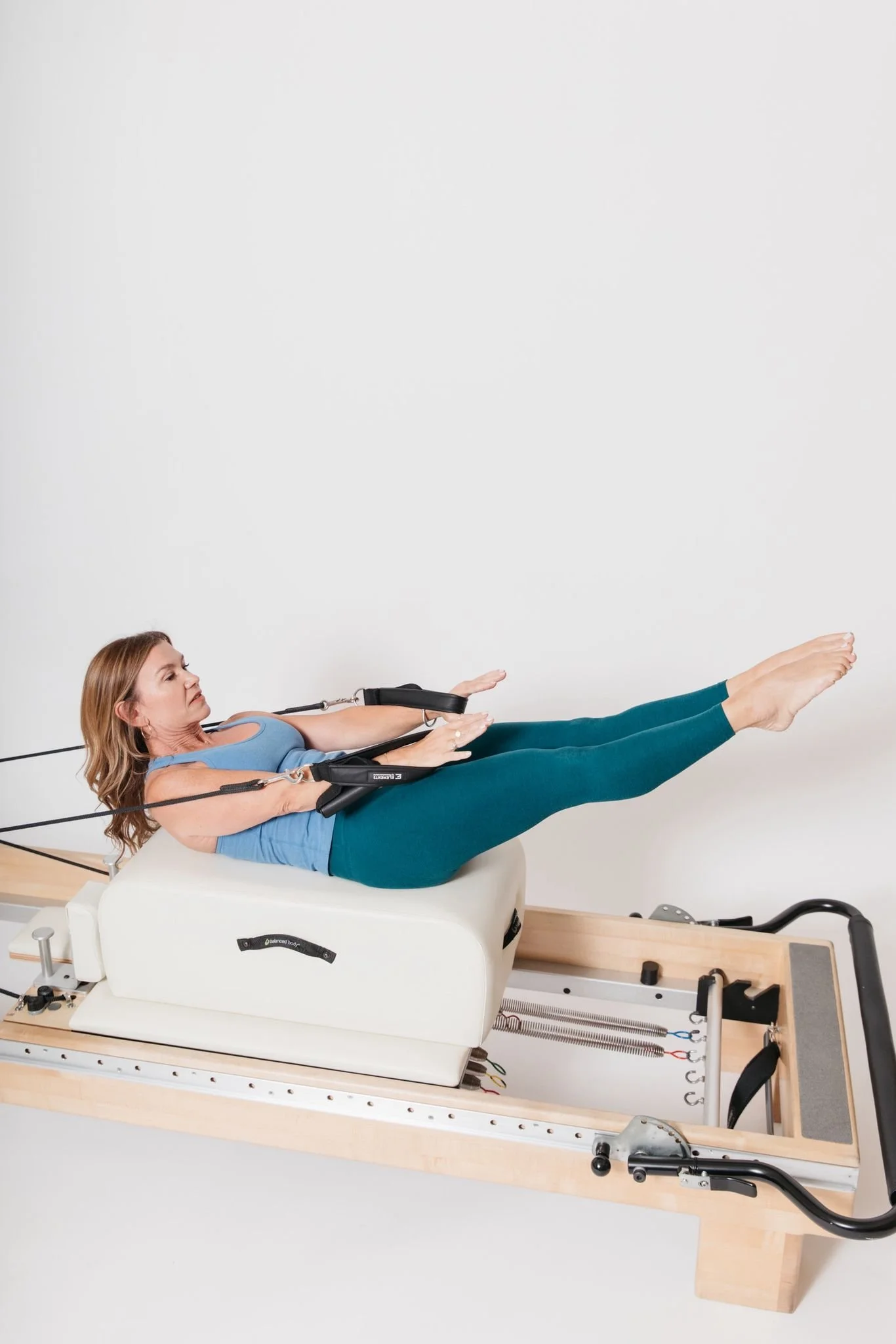 Woman exercises on a Pilates reformer machine, lying on her back with legs extended and arms outstretched, in a Pilates studio.