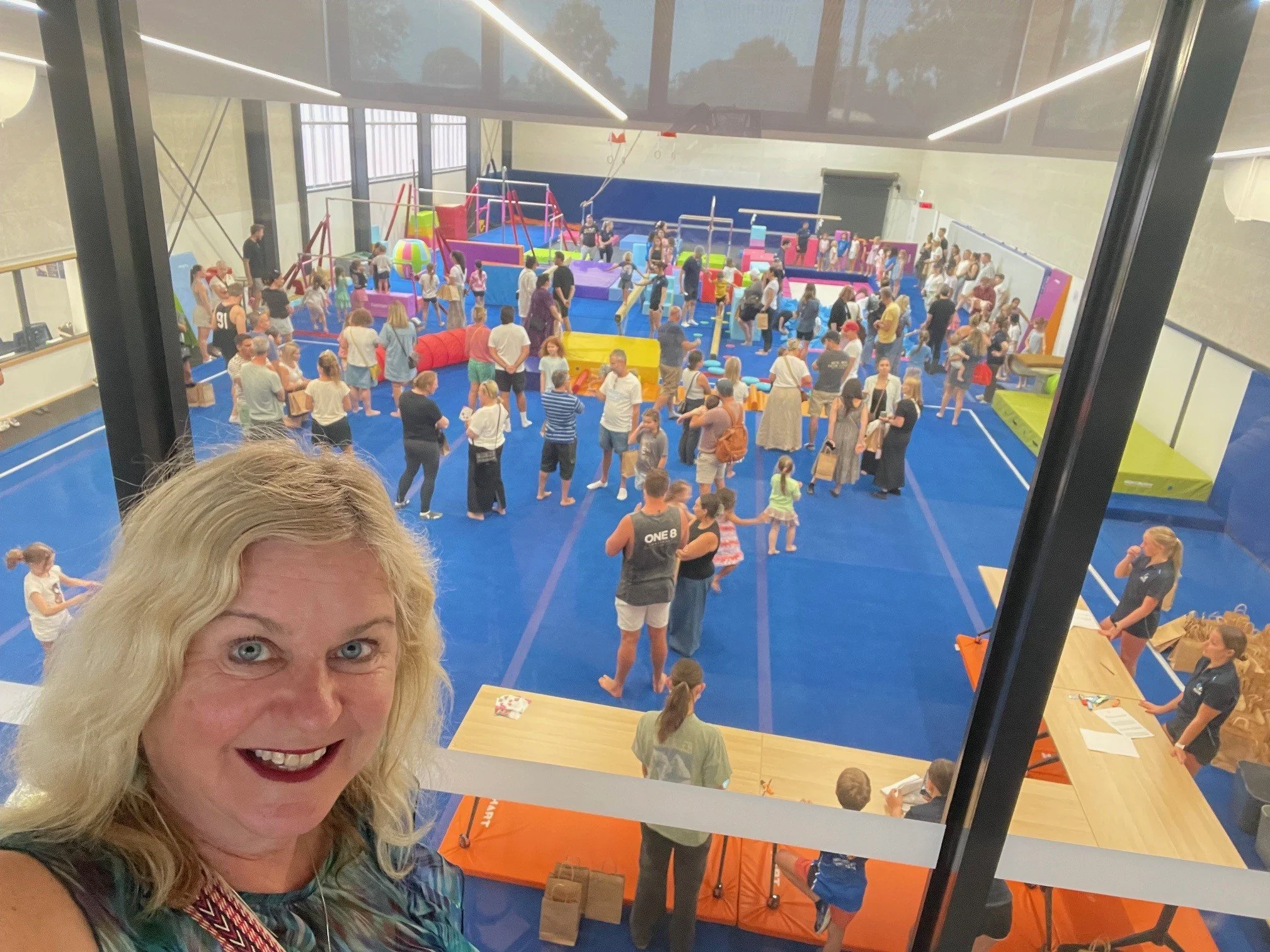 A woman taking a selfie in front of an indoor play gym filled with children and adults. The gym has blue padded flooring, colorful play structures, and a high ceiling with bright lighting.