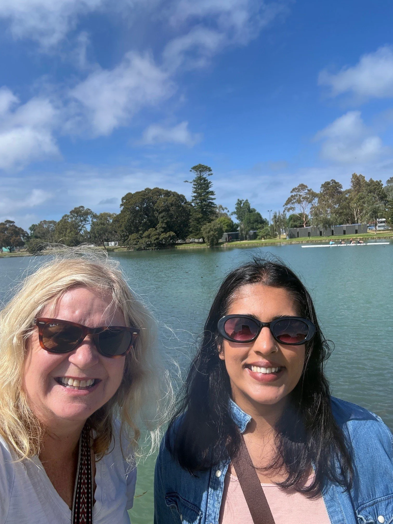 Two women Di McDonald and Sonal Sudan taking a selfie by a body of water outdoors on a sunny day, with trees and a boat in the background.