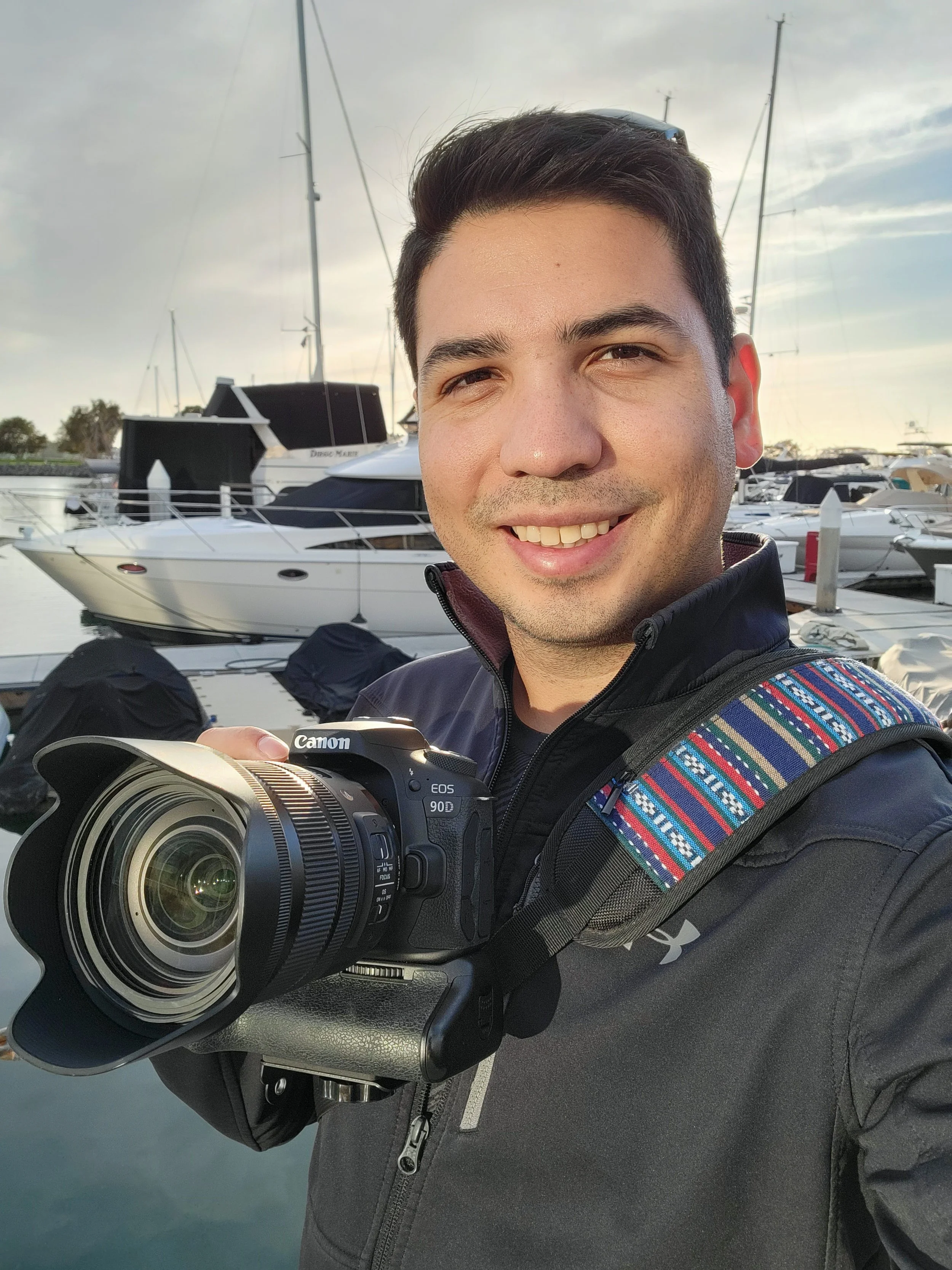 Jonathan smiling at the camera holding a Canon EOS 90D camera at a marina with boats and yachts in the background.