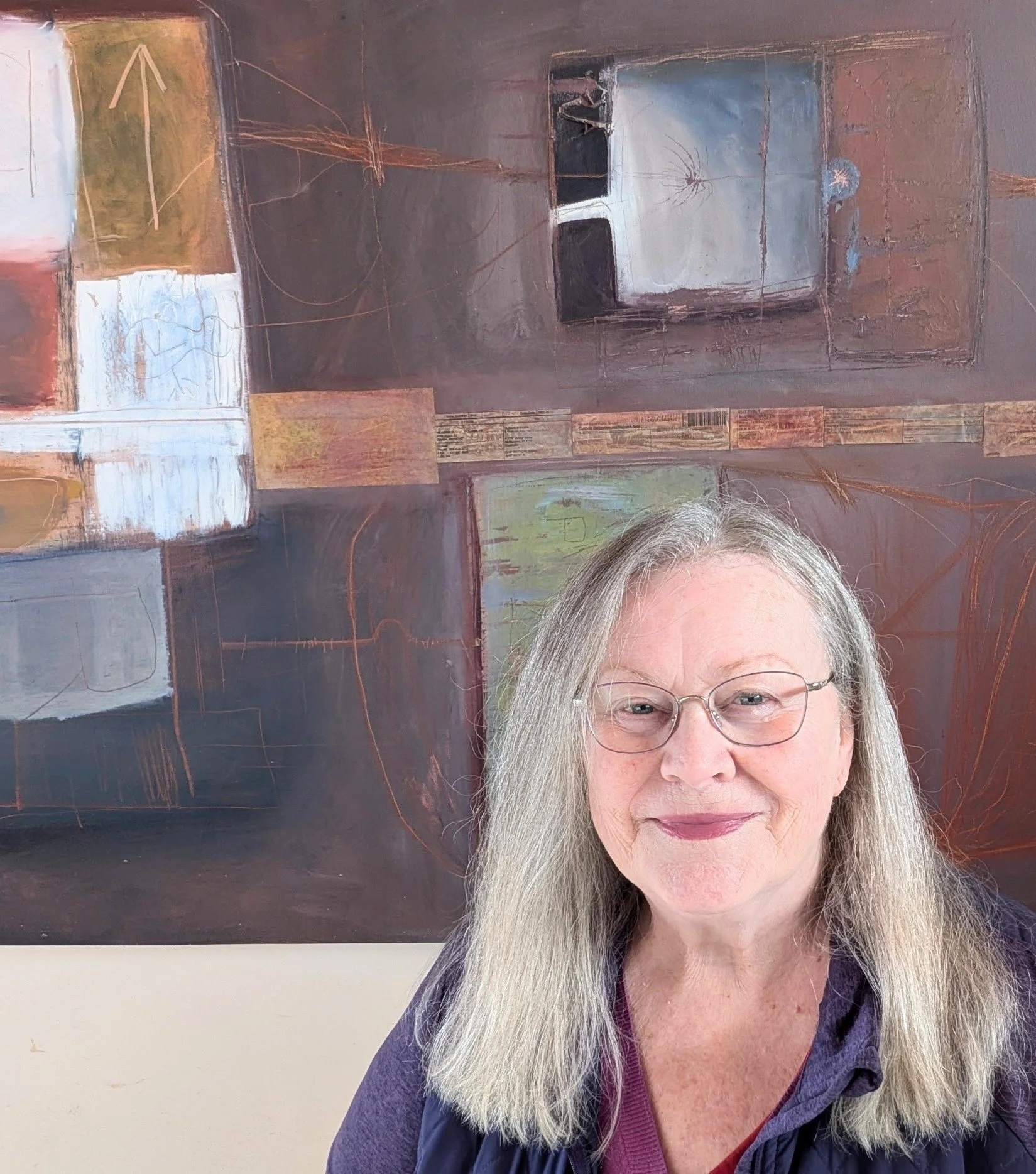 A woman with long hair in front of a wall with abstract paintings. She is smiling and wearing a dark jacket over a maroon top.