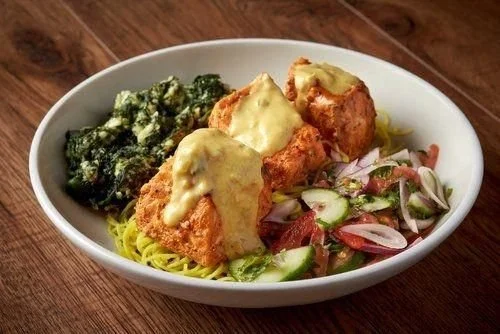 Bowl of pasta with fried chicken topped with melted cheese, side of sautéed greens, and a cucumber and tomato salad.