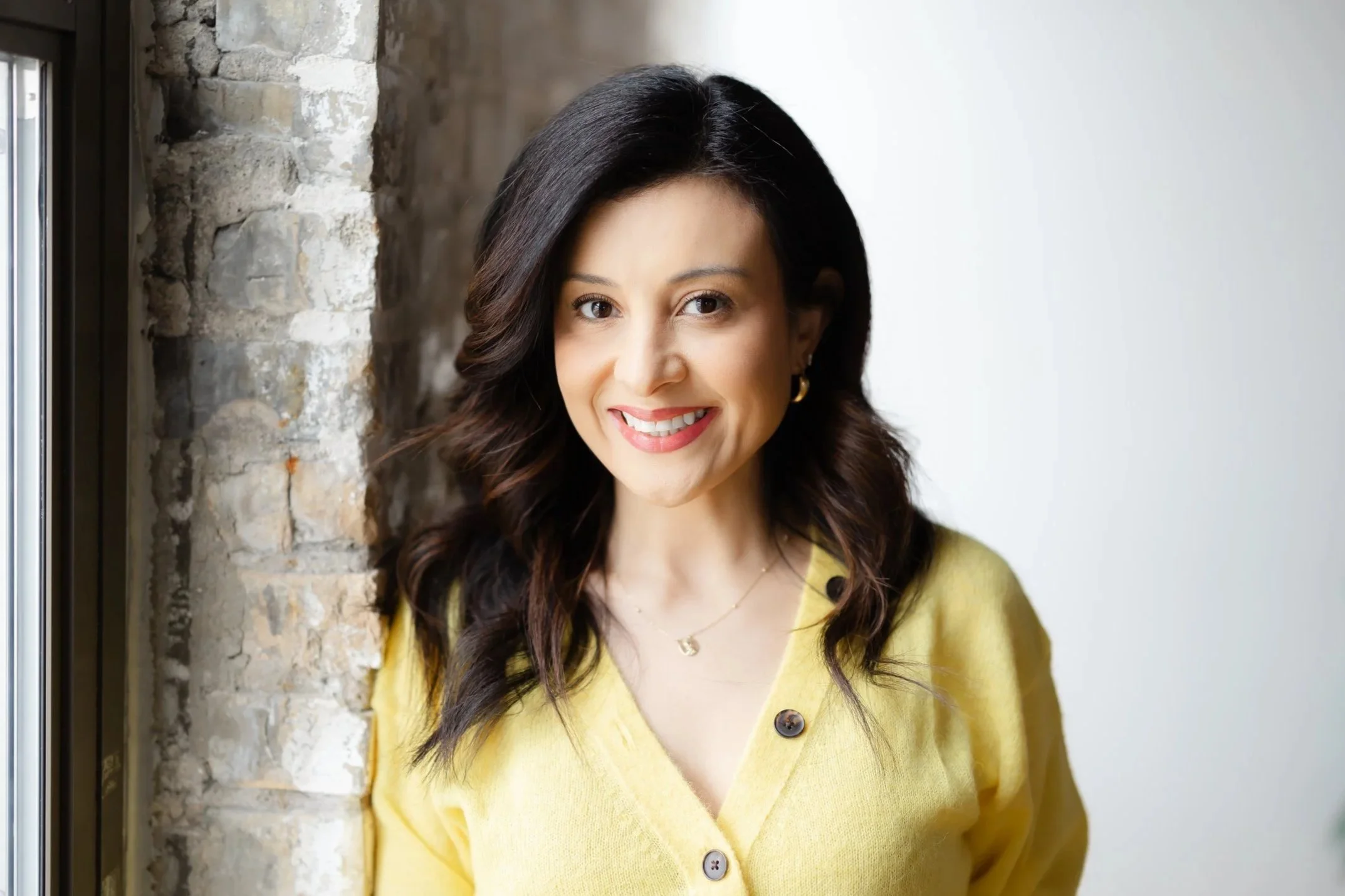Smiling woman in yellow sweater standing next to a window and brick wall.