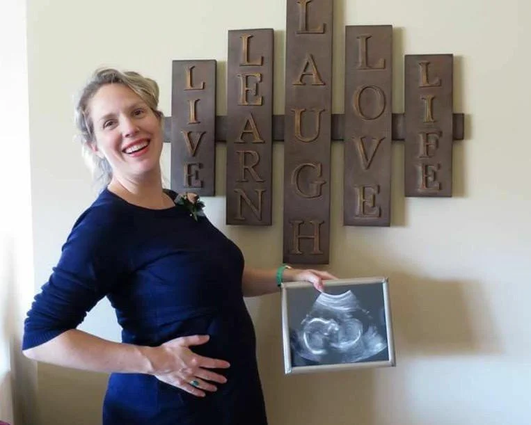 Pregnant woman holding ultrasound photo with wall art saying "Live, Learn, Laugh, Love, Life."