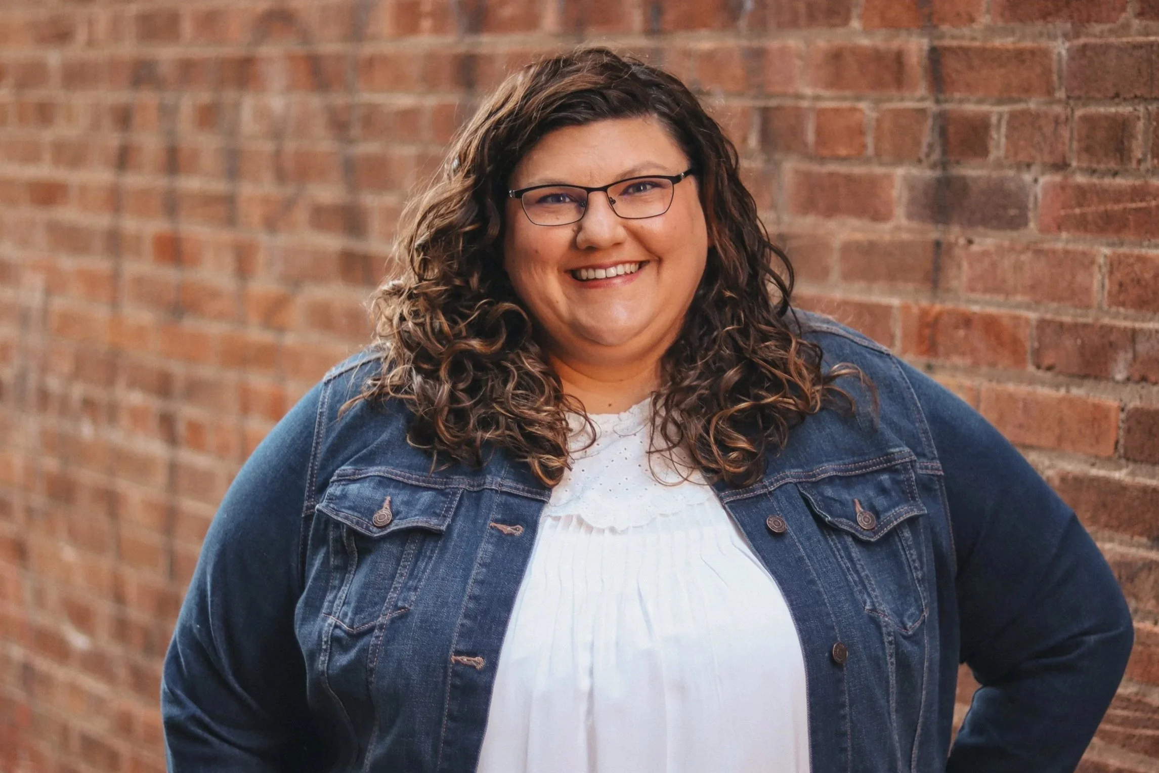 Woman smiling in front of a brick wall, wearing a denim jacket and glasses.