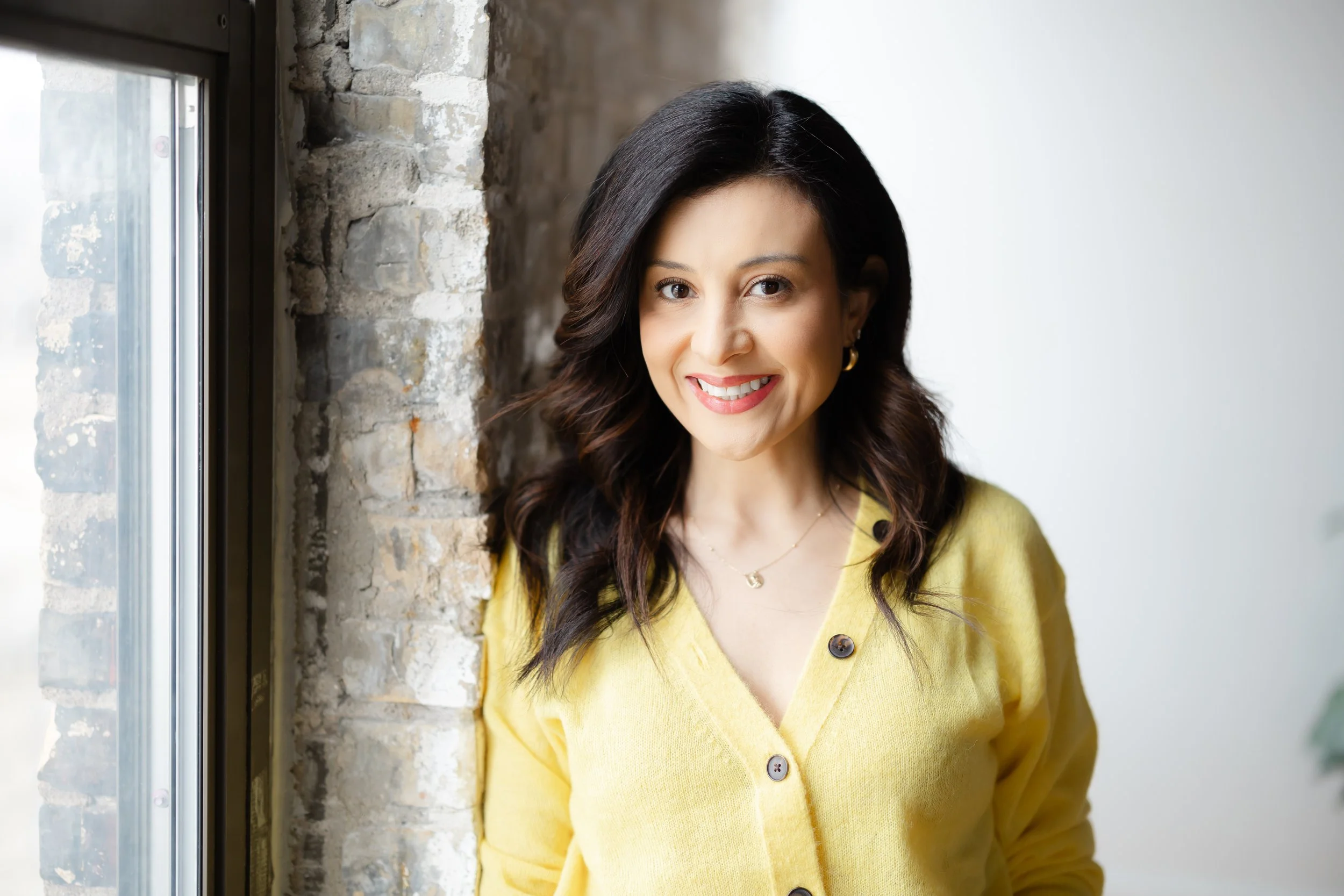 Smiling woman in yellow sweater standing next to a window and brick wall.