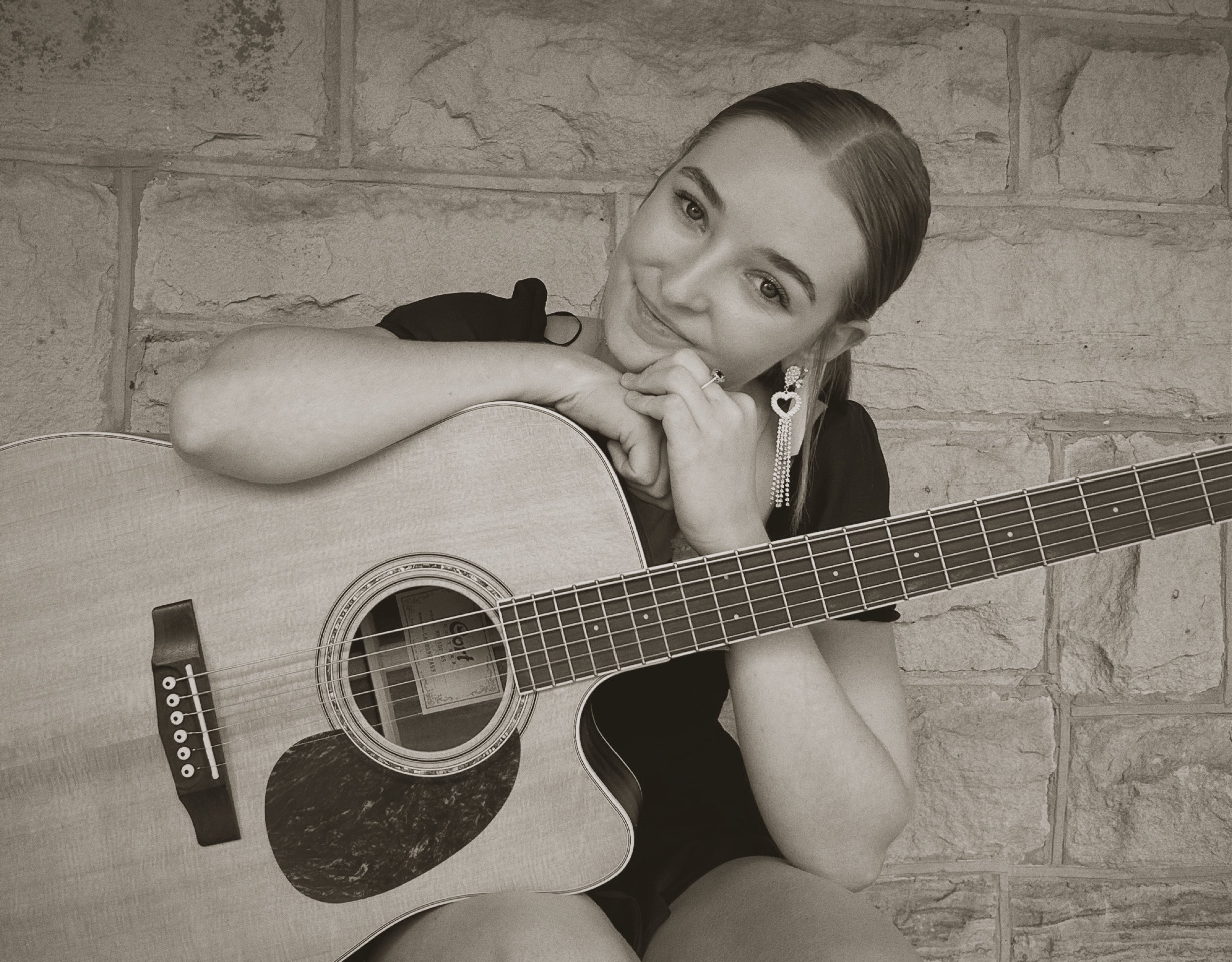 A young woman with long hair and earrings sitting against a brick wall, holding an acoustic guitar and smiling at the camera.