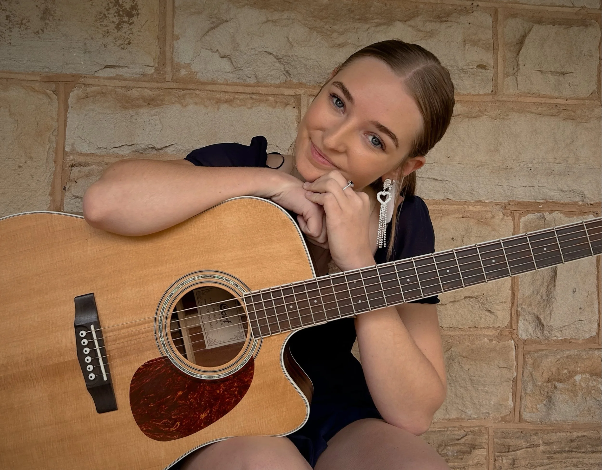 A young woman with blonde hair and blue eyes, wearing earrings and a dark top, is resting her chin on her hands, which are folded over the body of an acoustic guitar. She is sitting against a textured stone wall.