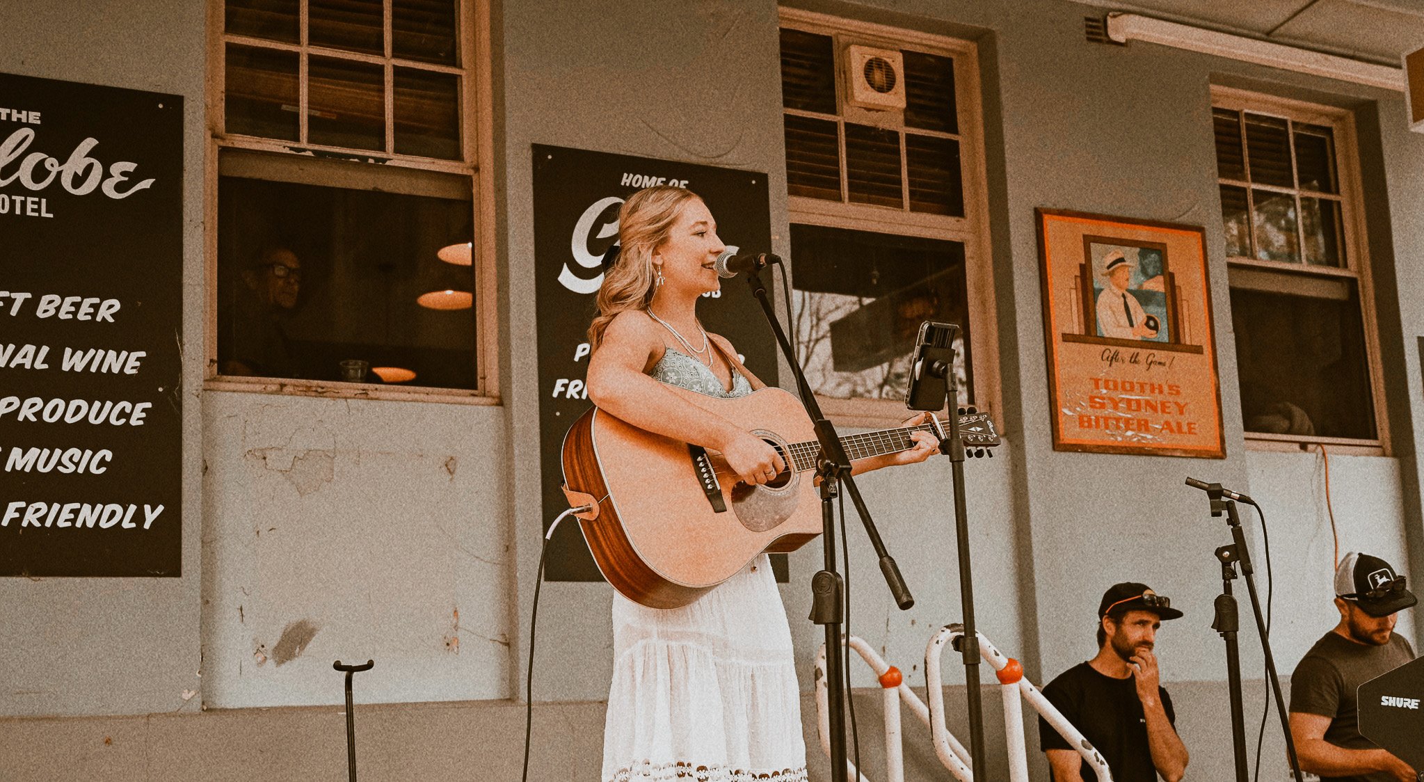 A woman with blonde hair singing and playing an acoustic guitar on outdoor stage, with two men sitting nearby, at a venue with signs and windows behind.