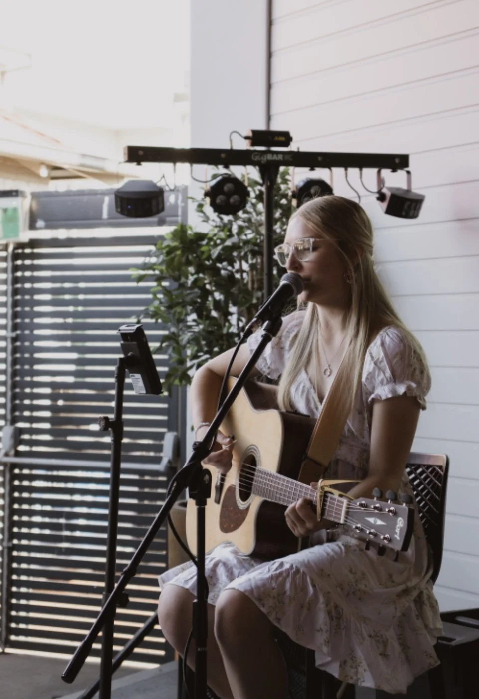 A woman with glasses singing into a microphone while playing an acoustic guitar at an indoor-outdoor venue.