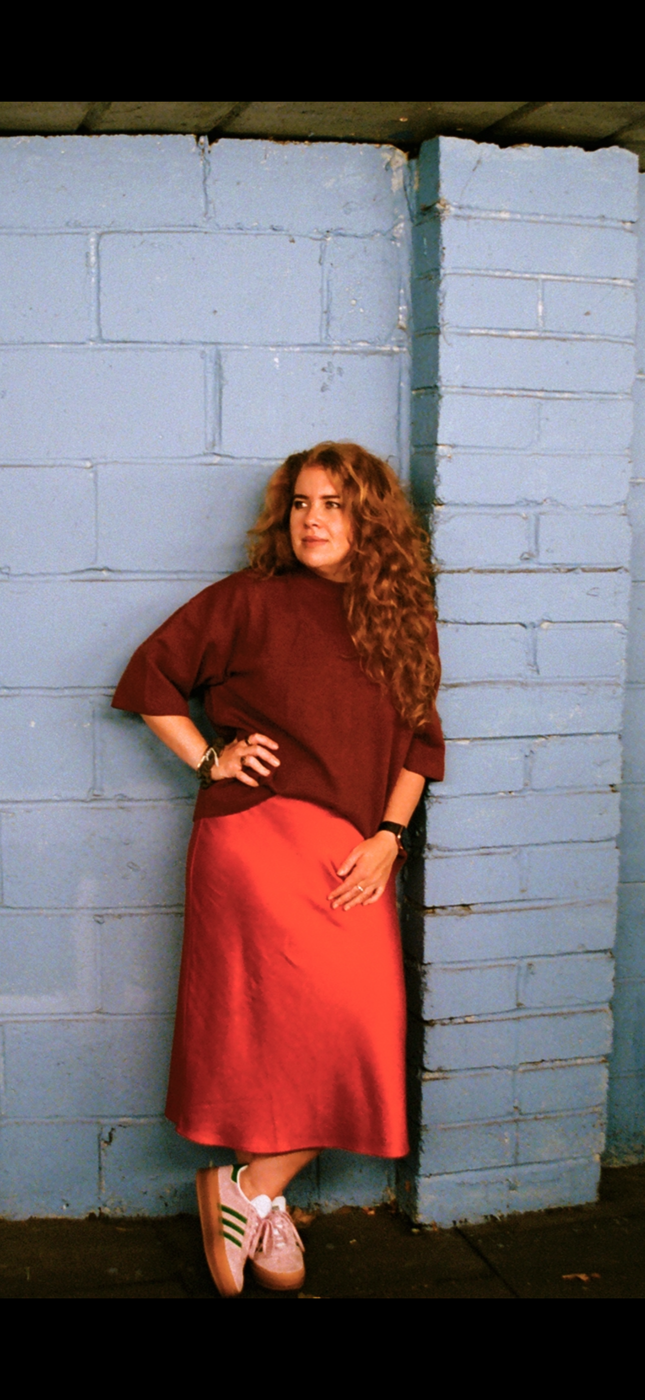 A woman with curly red hair wearing a maroon top and a long red skirt, standing with her hand on her hip against a blue painted brick wall, looking to her left.