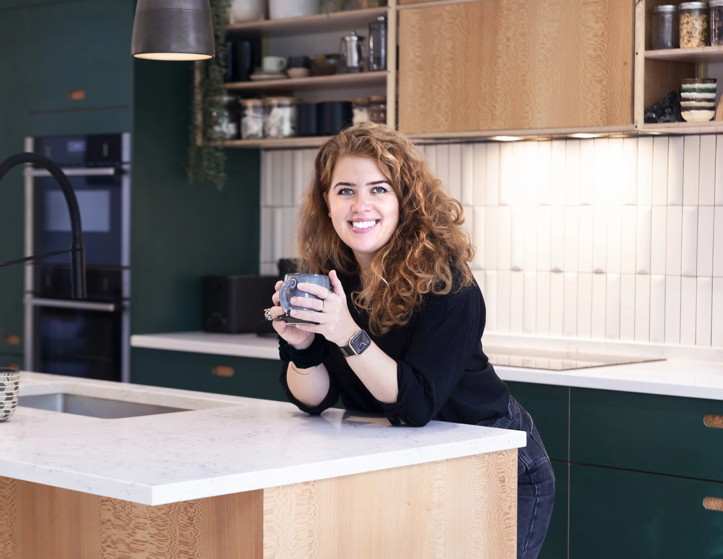 A woman with curly red hair smiling while holding a coffee cup in a modern kitchen with wooden and green cabinets.