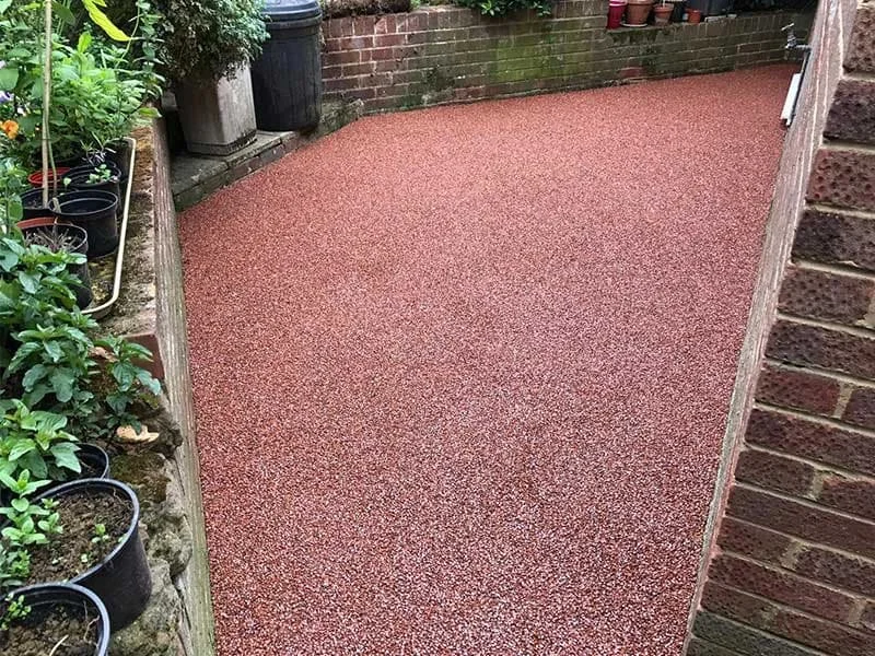 A small outdoor patio area with a red textured surface, bordered by a brick wall and a raised garden bed with plants and potted flowers.