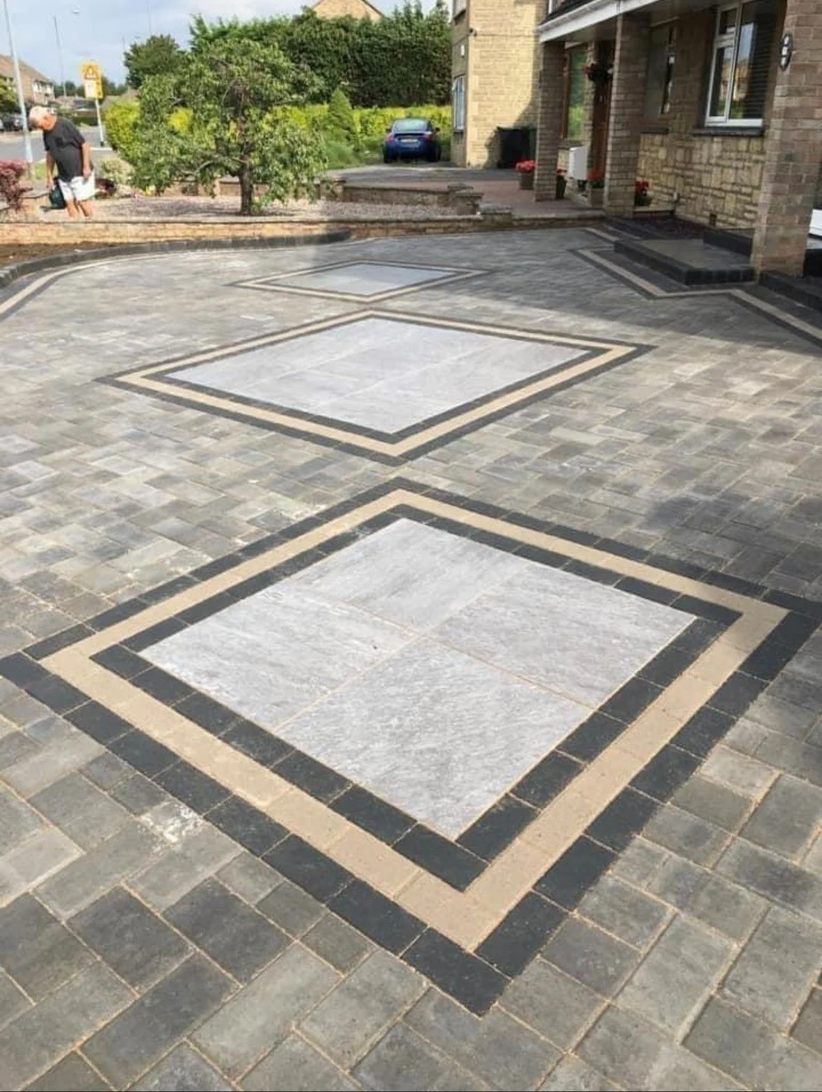 A driveway with decorative stone pattern, featuring large square stone tiles bordered by black and beige stones in geometric design.