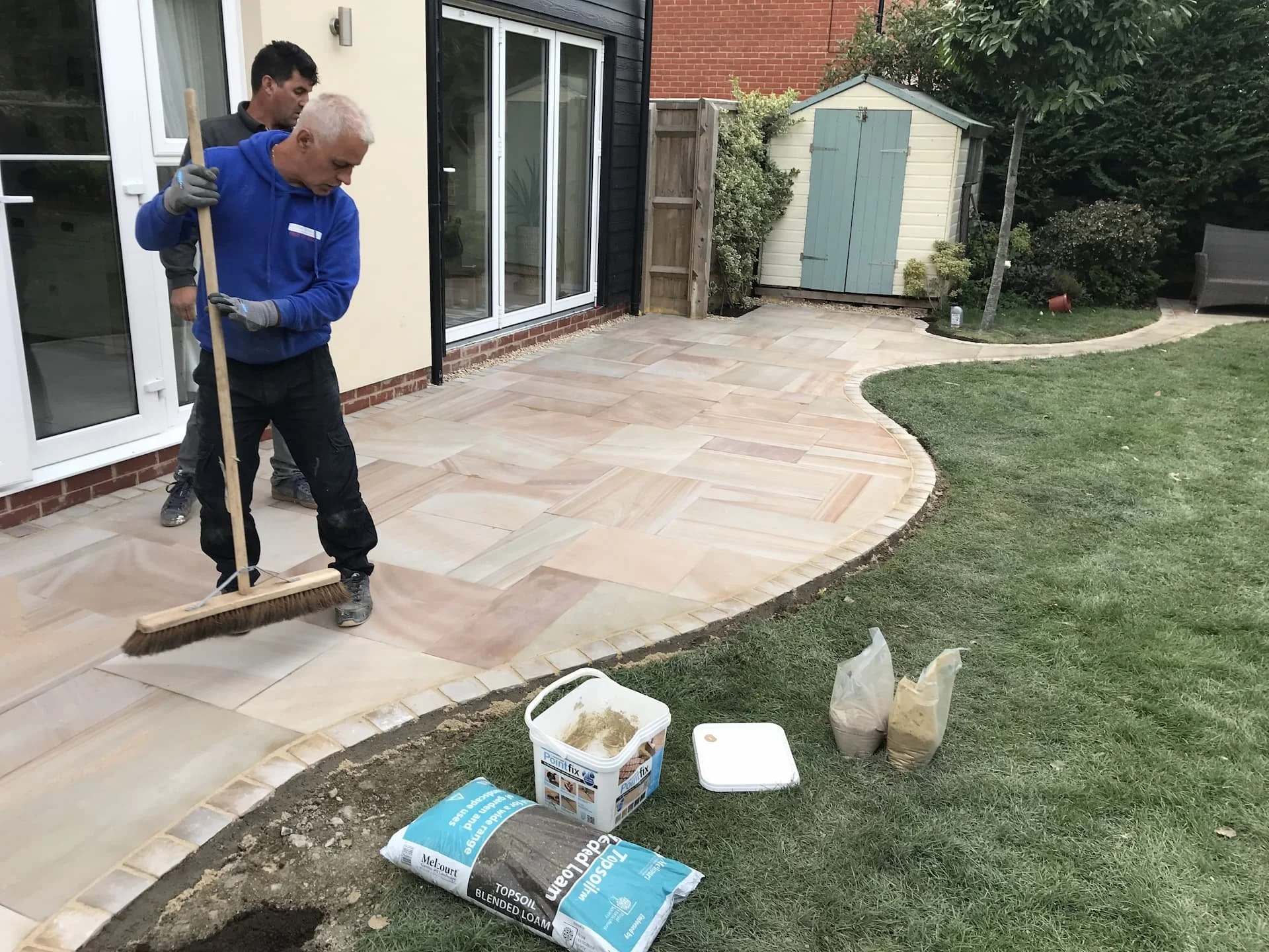 Two men working on laying a stone patio in a backyard, with one sweeping and the other guiding the placement of stones. There are gardening supplies, bags of sand or gravel, and a bucket in the foreground, with a garden shed and trees in the backgrou