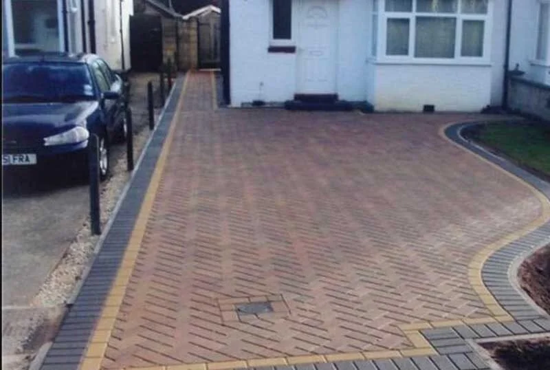 Newly paved brick driveway with a curved design, bordered by black and yellow edging, leading to the front door of a house, with parked cars on the side.
