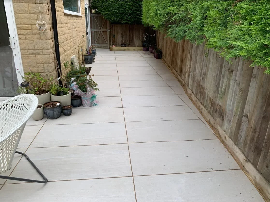 A backyard patio with potted plants along the house wall, a wooden fence on the right, and green trees overhead.