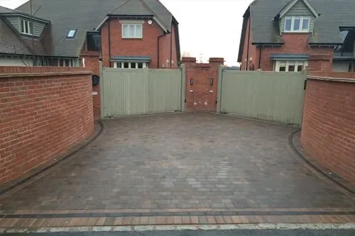 A paved driveway with brick walls on either side and a wooden gate at the end, leading to residential houses.