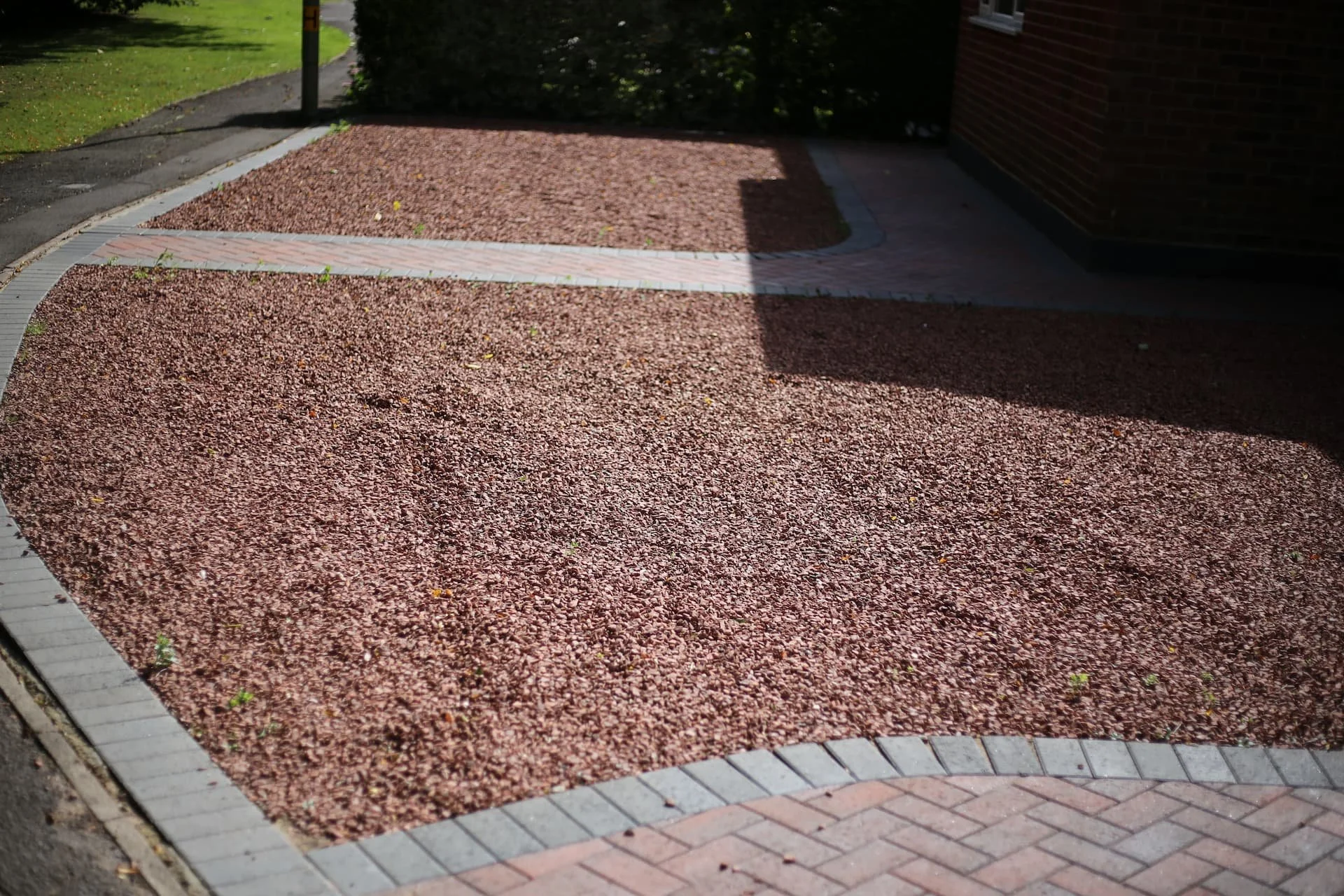 A sidewalk with a pinkish-red gravel surface bordered by concrete and bricks, next to a brick building and a grassy area.