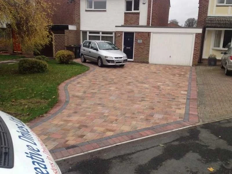 View of a house driveway with new brick pavers, a parked silver car, and a garage door. The driveway has a curved design with a border of darker bricks.