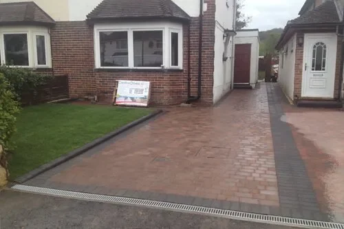 Image of a residential driveway with brick paving, a green lawn, and a house with large front windows. A sign is placed on the lawn near the house. The driveway is bordered by a dark gray strip along the edges.
