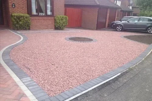 A neatly landscaped driveway with red gravel, gray brick border, and a circular drain in the center, in front of suburban houses.
