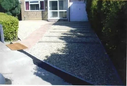 A driveway with a gravel section next to a concrete section, leading to a house with a garage. Shadows and bushes are visible.