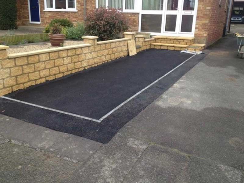 A designated parking space outlined with white lines on new asphalt in front of a house, with a low brick wall, potted plants, and steps leading to the entrance.
