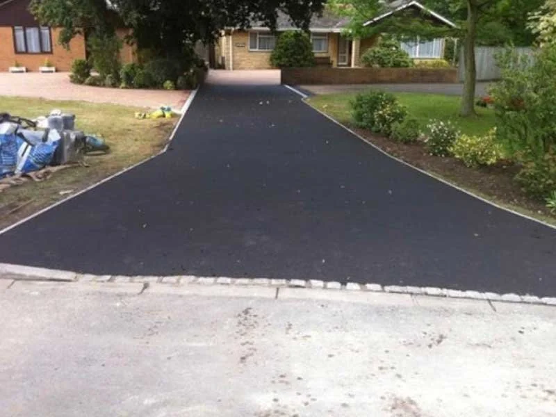 Freshly paved black driveway leading to a house with a small front porch, flanked by green bushes and trees.