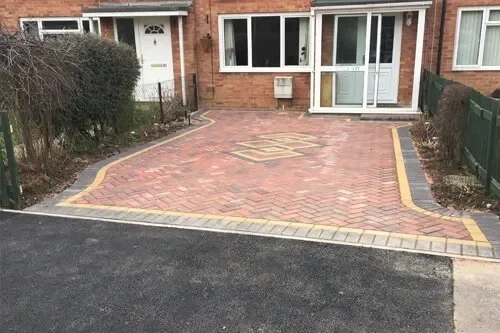 A paved driveway with a decorative border leading to a house with a porch and large windows, bordered by bushes and a fence.