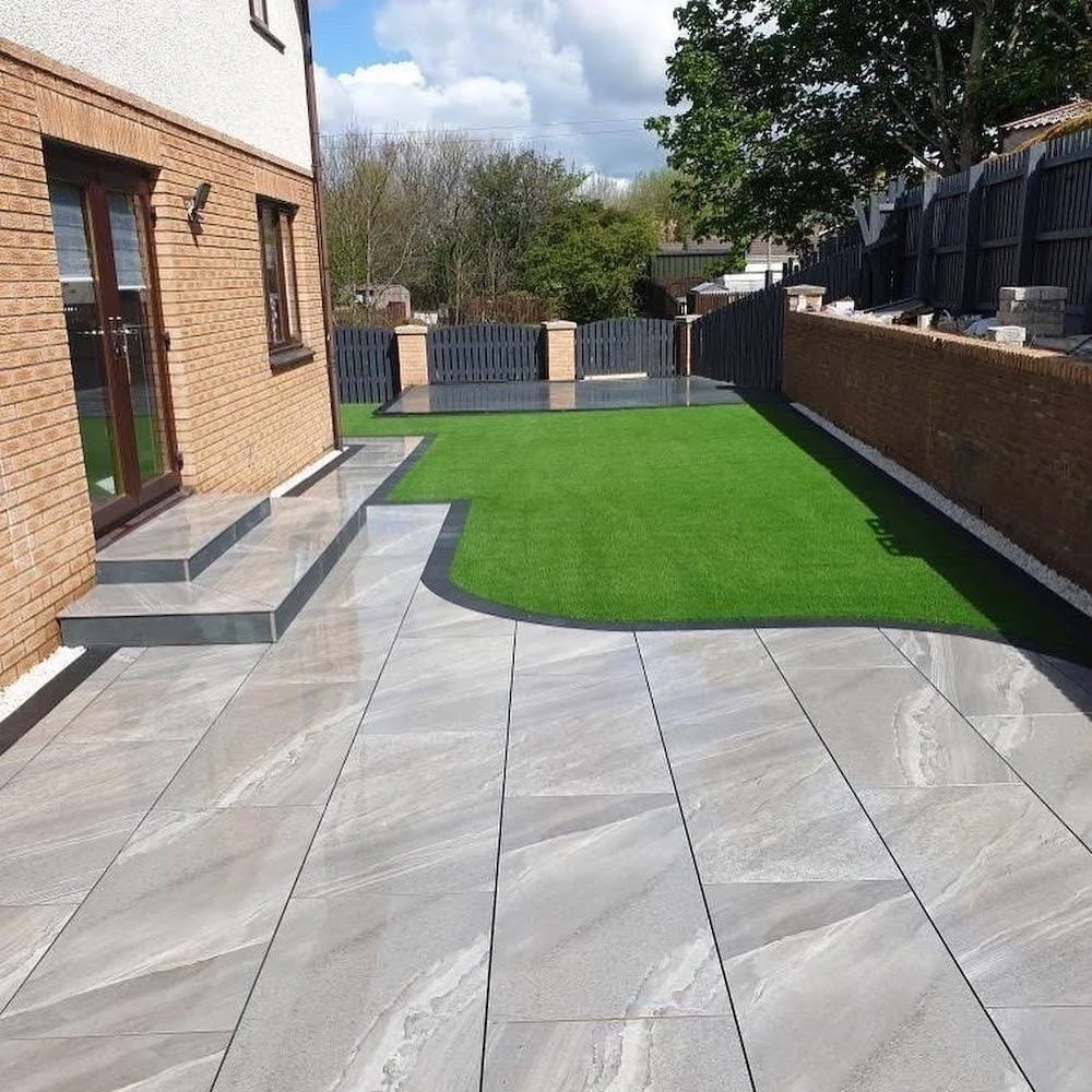 A backyard patio with light gray marble tiles, small steps leading up to a sliding glass door, and a patch of artificial green grass. There is a black fence and brick wall along the perimeter, with some trees and cloudy sky in the background.